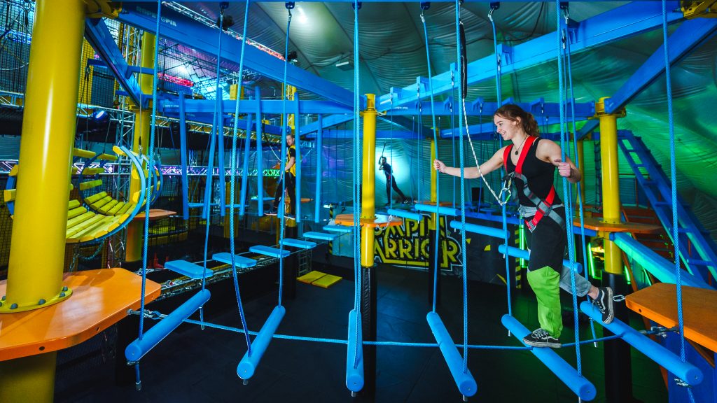 Junge Frau beim Klettern im Indoor-Hochseilgarten bei Jumpin Warrior title: Klettern im Indoor-Hochseilgarten bei Jumpin Warrior